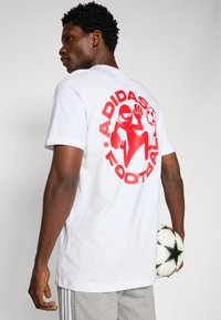 White cotton t-shirt featuring a large red graphic on the back with "ADIDAS FOOTBALL," and a playful cartoon character holding a soccer ball.