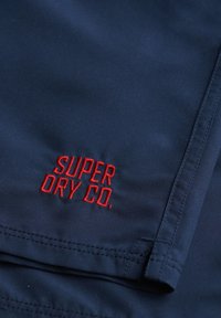 Pantalones cortos de tela azul marino con el texto rojo bordado "SUPER DRY CO." Textura suave con costuras visibles y detalle en el dobladillo.