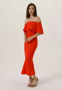 Piros, vállról lelógó ruha fodros nyakkivágással és testhez simuló szilhuettel, amely a szegélyenélél kiszélesedik. Bézs magassarkú szandálokkal viselve.