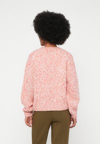 Persona con cabello rizado, vistiendo un suéter de punto rosa y blanco y pantalones de color verde oliva, de pie mirando hacia atrás contra un fondo liso.