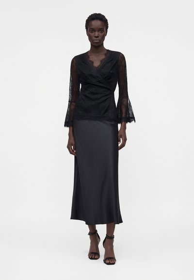 Γυναίκα στέκεται σε λευκό φόντο, φοράει μαύρο δαντελένιο τοπ με μακριά μανίκια, μαύρη σατέν μίντι φούστα και μαύρες γόβες με λουράκι στον αστράγαλο.