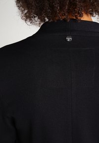 Schwarzes Strickkleid mit rundem Ausschnitt, einem kleinen silbernen Knopf und einem quadratischen Aufnäher auf der Rückseite. Weiche Textur mit minimalen Details.