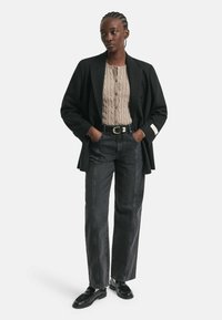 Schwarzer taillierter Blazer, beiger strukturierter Knopfpullover, hoch geschnittene graue Jeans mit weißen Nähten und schwarze Schuhe.