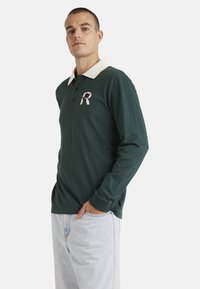 Long-sleeve polo shirt in dark green cotton with a cream collar, featuring a embroidered logo on the chest and ribbed cuffs.