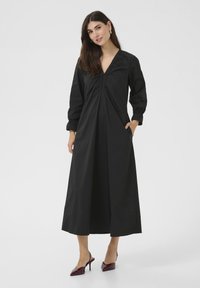 Must mustriga kleit V-kaelusega, kogutud esiosa detaile ja küljetaskutega. Sile kanga, voolav siluett, kombineeritud burgundi värvi kontsakingadega.