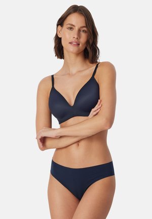 Donna con capelli corti e castani che indossa un completo intimo nero abbinato, in piedi con le braccia incrociate davanti a uno sfondo neutro.