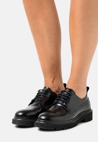 Černé leštěné kožené šněrovací boty s brogue detaily nošené na bosých nohách na světlém pozadí.