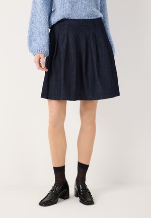 Frau trägt einen plissierten dunklen Jeansrock, hellblauen Strickpullover, schwarze durchsichtige Socken und schwarze Loafers mit Absatz und steht vor einem einfarbigen Hintergrund.