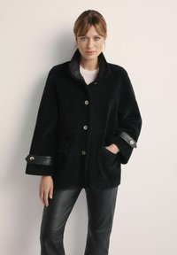 Czarna futrzana peleryna z wysokim kołnierzem, srebrnymi guzikami i skórzanymi akcentami na mankietach. Widoczne kieszenie; model nosi czarne skórzane spodnie.