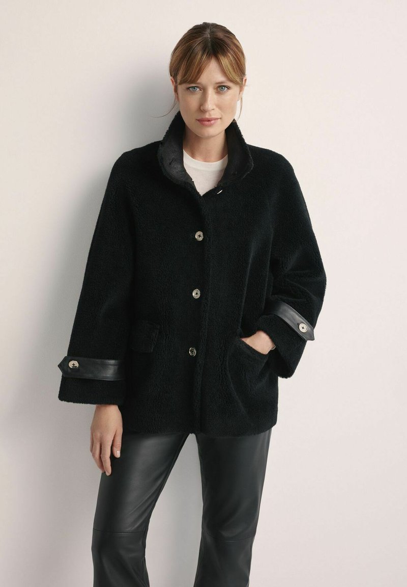 Czarna futrzana peleryna z wysokim kołnierzem, srebrnymi guzikami i skórzanymi akcentami na mankietach. Widoczne kieszenie; model nosi czarne skórzane spodnie.