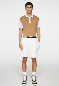 Beżowa i biała koszulka polo z krótkimi rękawami, z logo na piersi, zestawiona z białymi szortami i czarnymi sneakersami. Gładki, sportowy fason.