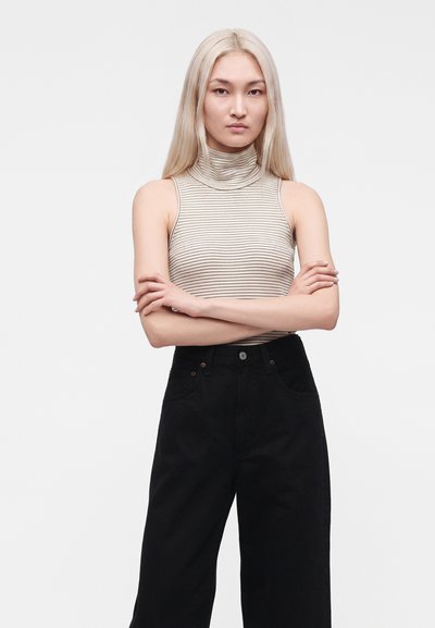 Jeune femme aux longs cheveux blonds portant un col roulé sans manches beige rayé et un pantalon noir taille haute, les bras croisés, expression neutre.