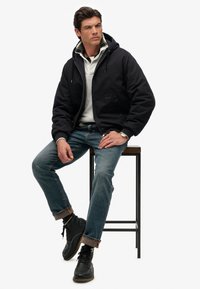 Zwarte capuchonjack, witte trui, blauwe jeans en zwarte laarzen. Inclusief een geruite kraagdetail, zittend op een kruk tegen een effen achtergrond.