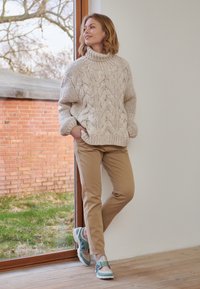 Cream cable-knit sweater with high collar, loose fit, paired with tan chinos and blue/green sneakers, set against a natural backdrop.