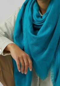 Personne portant une grande écharpe turquoise à motifs sur une veste beige, la main posée sur un genou plié.