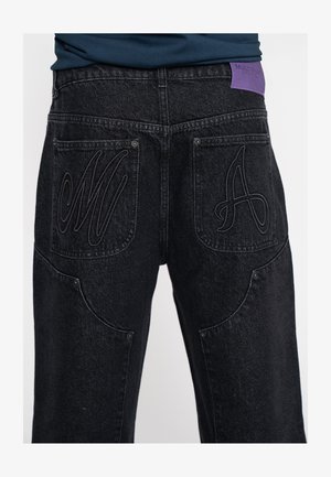 I jeans in denim neri presentano due tasche posteriori con lettere ricamate alzate "M" e "A". Un'etichetta del marchio di colore viola è cucita sul cinturino.