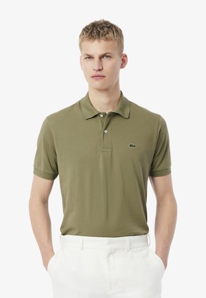 Olivgrön polo med klassisk krage, korta ärmar och broderad logotyp på bröstet, i kombination med vita byxor.