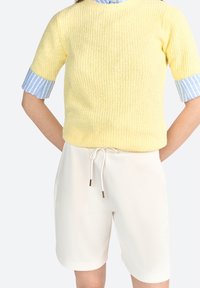 Geel gebreide trui met korte mouwen en een blauwe gestreepte manchette, gecombineerd met crèmekleurige shorts met een trekkoord in de taille en gouden uiteinden.