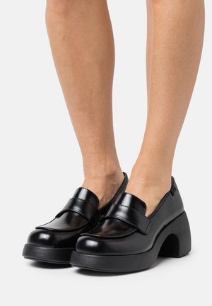 Zwarte glanzende loafers met een gebogen hoge hak, met een soepele textuur en twee brede banden over de bovenkant. Instapontwerp.