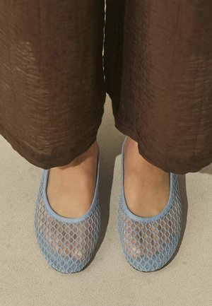 Feet wearing light blue mesh slip-on shoes paired with loose brown pants on a beige surface.