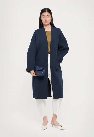 Jūras zila garā jaka ar šallītes apkakli, apvienota ar zelta topu, baltām biksēm un teksturētu zilu maciņu, ko papildina krēmkrāsas mūlas.