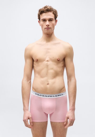 Νέος άνδρας όρθιος, φορώντας ανοιχτό ροζ boxer briefs Polo Ralph Lauren, γυμνός από το στήθος, σε απλό ανοιχτό φόντο.