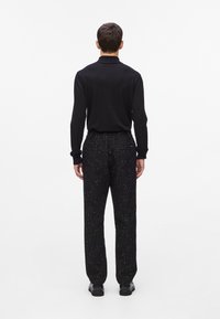 Fekete hosszúujjú ing, slim fit fazonban, texturált fekete nadrággal, amelyen finom minta található, egyenes lábakkal, fekete cipővel viselve.
