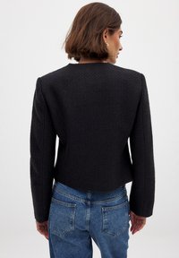 Giacca nera texture con una silhouette cropped, spalle strutturate e maniche lunghe. Indossata con jeans blu a vita alta.