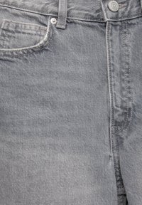 Gros plan sur un jean en denim gris délavé montrant la poche avant, la boucle de ceinture, les détails de couture et le bouton métallique de fixation.