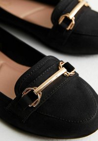 Zwarte suède loafers met een ronde neus, voorzien van een gouden metalen accent en stiksels op de bovenkant. Platte zool.