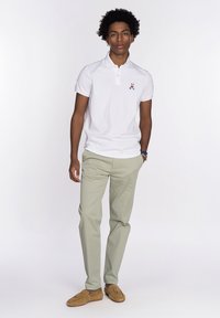 Camisa polo blanca con un pequeño diseño bordado, combinada con pantalones verde claro. Los accesorios incluyen una pulsera multicolor y mocasines marrones.
