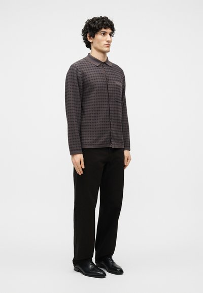 Άνδρας που στέκεται, φορώντας μακρυμάνικο πουκάμισο με σχέδια και φερμουάρ, μαύρο παντελόνι και μαύρα επίσημα παπούτσια, μπροστά σε απλό λευκό φόντο.