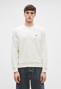 Bílý svetr s kulatým výstřihem, žebrovanými manžetami a spodním lemem. Na hrudi je malá červená značka. Nosit s tmavým denimem, který ukazuje uvolněný střih.