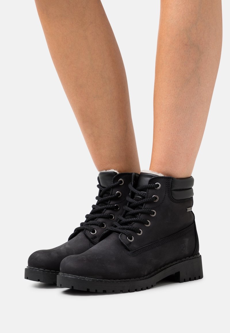 Bottines noires à lacets avec col rembourré et semelles épaisses portées sur des jambes nues sur un fond clair uni.