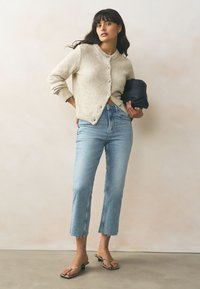 Krémový pletený kardigan s kulatým výstřihem a zapínáním na knoflíky, doplněný světle modrými zkrácenými džíny a sandály na podpatku na tenkých řemíncích. V ruce černé psaníčko.