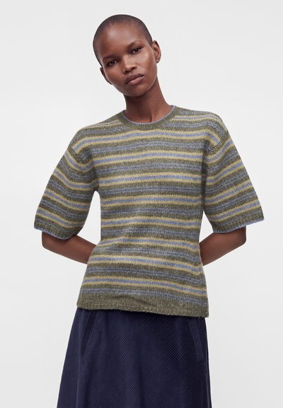 Personne aux cheveux courts portant un pull à manches courtes rayé vert, bleu et jaune et une jupe bleu foncé sur un fond uni.