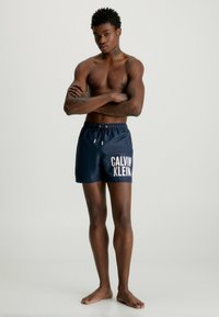 Shorts da piscina blu navy con vita elastica e cordino, con stampa del logo "CALVIN KLEIN" in bianco sulla parte anteriore sinistra. Tessuto dalla texture liscia.