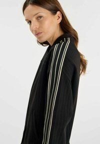 Svart jacka med vertikala vita ränder, med dragkedjestängning, sidofickor och en avslappnad passform. Texturerad tyg med sportiga detaljer.