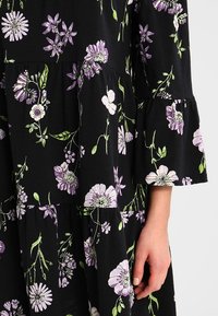 Czarna sukienka w kwiaty z fioletowymi i białymi motywami, długimi rękawami, warstwowym krojem i miękką fakturą. Ręka widoczna na dole.