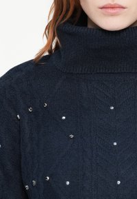 Pull en tricot bleu marine avec col roulé, orné d'un motif en torsades et de sequins décoratifs éparpillés, avec un tissu texturé et un col côtelé.