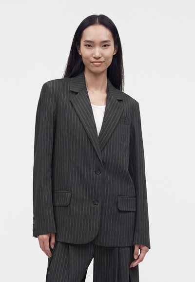 Personne aux longs cheveux foncés porte un costume sombre à rayures et un haut blanc, debout devant un fond clair uni, tournée vers l'avant et souriante.
