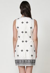 Ärmelloses weißes Kleid mit schwarzer geometrischer Stickerei, engen Silhouette und gemustertem Saum, aus leichtem Stoff gefertigt.