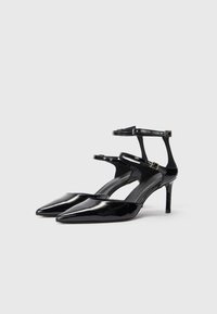 Črne lakirane usnjene pete s koničastimi prsti, trakovi okrog gležnja in tankim stiletto peto. Gladka tekstura in minimalistična zasnova.