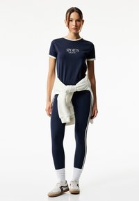 Marineblå atletisk t-shirt med hvid kant og "SPORTS" tekst, parret med tætsiddende marineblå leggings med sidelister og hvide sneakers.