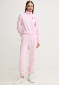 Tuta Adidas rosa chiaro con colletto alto e zip, dotato di vita e polsini elastici, abbinato a sneakers bianche.