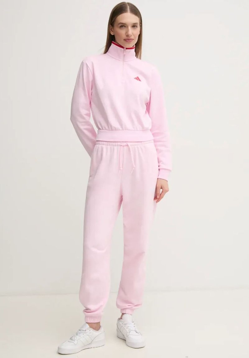 Tuta Adidas rosa chiaro con colletto alto e zip, dotato di vita e polsini elastici, abbinato a sneakers bianche.
