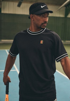 T-shirt de sport noir avec des accents blancs, à manches courtes, col rond, en tissu texturé, et logo sur la poitrine gauche. Présenté avec une raquette de tennis.