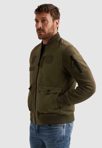 Muž ve vojensky zelené bomber bundě s nášivkami a kapsami na zip, v modrých džínách, otočený doleva s rukama v kapse.