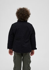 Juodas medvilnės švarkas su stovinčiu aptaku ir sagomis prie rankovių, derinamas su alyvuogių žalios spalvos cargo kelnėmis, turinčiomis daugybę kišenių.