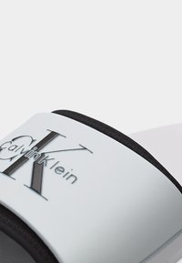 Λευκό λουράκι σανδάλιας με ανάγλυφο μαύρο λογότυπο "Calvin Klein" πάνω σε υφασμένο μαύρο πέλμα, με λευκό φόντο.